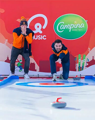 Twee mannen zijn bezig met curling op een ijsbaan met op de achtergrond logo's van Qmusic en Campina
